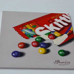 Op de afbeelding: Een schilderij van een rode, witte en gele Skittles-zak met het woord "original" erop. De zak is open en er vallen Skittles uit. Er liggen zes Skittles op een grijze achtergrond. De Skittles zijn rood, oranje, geel, groen en paars. De handtekening van de kunstenaar staat in de rechterbenedenhoek.