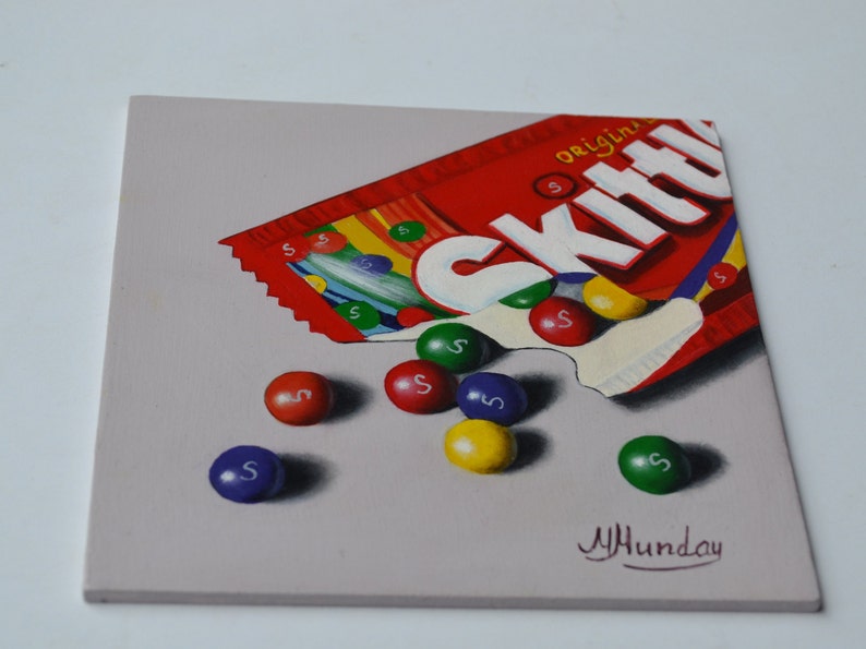 Op de afbeelding: Een schilderij van een rode, witte en gele Skittles-zak met het woord "original" erop. De zak is open en er vallen Skittles uit. Er liggen zes Skittles op een grijze achtergrond. De Skittles zijn rood, oranje, geel, groen en paars. De handtekening van de kunstenaar staat in de rechterbenedenhoek.
