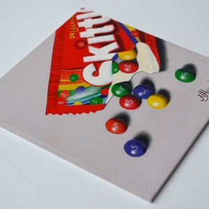 Op de afbeelding: Een schilderij van een gedeeltelijk geopende zak Skittles snoep met het woord "Skittles" erop gedrukt. De zak is rood en wit met een regenboog van kleuren aan de onderkant. Verschillende Skittles snoepjes vallen uit de zak en op een grijs oppervlak. De snoepjes zijn rood, oranje, geel, groen en paars. De handtekening van de kunstenaar, "J. Hummer", staat in de rechterbenedenhoek van het schilderij.