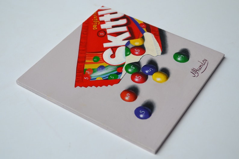 Op de afbeelding: Een schilderij van een gedeeltelijk geopende zak Skittles snoep met het woord "Skittles" erop gedrukt. De zak is rood en wit met een regenboog van kleuren aan de onderkant. Verschillende Skittles snoepjes vallen uit de zak en op een grijs oppervlak. De snoepjes zijn rood, oranje, geel, groen en paars. De handtekening van de kunstenaar, "J. Hummer", staat in de rechterbenedenhoek van het schilderij.