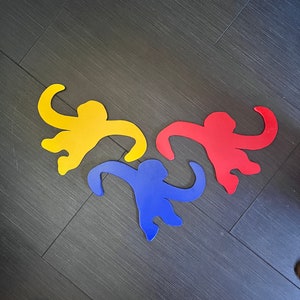 May include: Three cutout monkey shapes in yellow, red, and blue. The monkeys are arranged in a triangle shape on a dark gray surface.