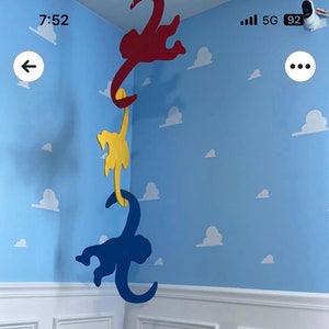 Op de afbeelding: Drie houten aap-uitsnijdingen die aan het plafond hangen. De bovenste aap is rood, de middelste aap is geel en de onderste aap is blauw. De apen hangen in een hoek van een kamer met een blauwe muur en witte wolken die op de muur zijn geschilderd.