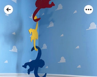 Puede incluir: Tres recortes de monos de madera colgando del techo. El mono de arriba es rojo, el mono del medio es amarillo y el mono de abajo es azul. Los monos están colgando en una esquina de una habitación con una pared azul y nubes blancas pintadas en la pared.