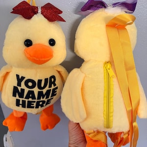 May include: Two plush yellow duck backpacks with orange beaks and feet. One has a red bow with a rhinestone center, the other a purple bow. The text "YOUR NAME HERE" is printed on the front of one duck. Each has a yellow zipper and ribbon straps.