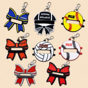 May include: A collection of eight silicone keychain holders for hair ties, featuring various shapes including bows, footballs, softballs, and volleyballs. Each item includes a metal lobster clasp and personalized names like Cameron, Elizabeth, Julie, Ava, and Lexie.