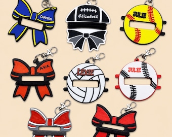 May include: A collection of eight silicone keychain holders for hair ties, featuring various shapes including bows, footballs, softballs, and volleyballs. Each item includes a metal lobster clasp and personalized names like Cameron, Elizabeth, Julie, Ava, and Lexie.