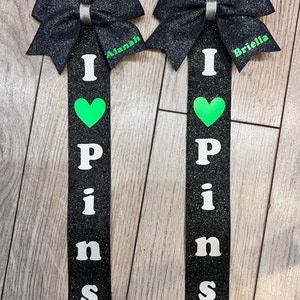 May include: Two black and silver glitter cheer bows with green hearts and the words "I love pins" on black ribbon. The bows are personalized with the names "Alanah" and "Briella".