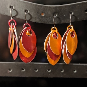 May include: Four pairs of handmade earrings with a scale-like design. The earrings feature a layered pattern of red, orange, and gold metal scales. Each pair hangs from a black hook. The earrings are displayed on a black stand.