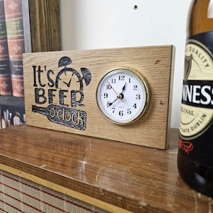Pode incluir: Um relógio de madeira com a frase "It's Beer O'Clock" gravada. O relógio tem um mostrador redondo com uma moldura dourada. Uma garrafa de Guinness é parcialmente visível no fundo.