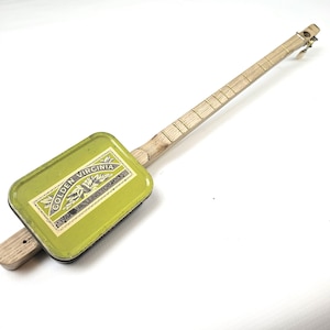 May include: A handmade cigar box guitar. The body is a light green rectangle with the text "GOLDEN VIRGINIA HAND ROLLING TOBACCO". The neck and fretboard are made of light-coloured wood.