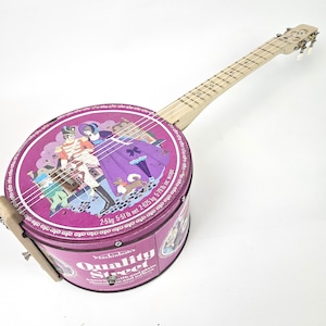 May include: A handmade banjo with a round body made from a Mackintosh's Quality Street candy tin. The tin is magenta with a colorful illustration. The neck is light wood. The banjo has four strings and tuning pegs.