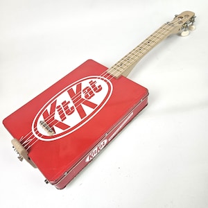 May include: A handmade cigar box guitar with a red Kit Kat candy box as the body. The guitar has a wooden neck, fretboard, and headstock with white tuning pegs. The Kit Kat logo is prominently displayed on the front.