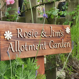 Könnte beinhalten: Ein Holzschild mit dem Text "Rosie & Jim's Allotment Garden" in weißen Buchstaben. Das Schild ist mit zwei weißen Holzblumen verziert.