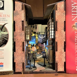 Puede incluir: Un edificio de ladrillo en miniatura con una puerta iluminada y un letrero que dice "Flourish & Blotts". El edificio está hecho de madera y pintado para que parezca ladrillo. El edificio está flanqueado por dos lomos de libros, uno para "El Señor de los Anillos" de J.R.R. Tolkien y el otro para "Dreamsongs" de George R.R. Martin.