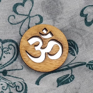 Può includere: Un ornamento rotondo in legno con un simbolo Om bianco intagliato. L'ornamento è su uno sfondo di tessuto con un motivo floreale.