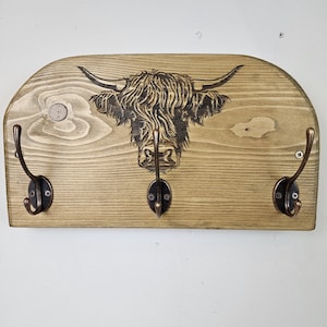 May include: Wooden wall-mounted coat rack with a Highland cow design. The light brown wooden board features three dark bronze hooks and a detailed engraving of a Highland cow's head. The rack is mounted with screws.