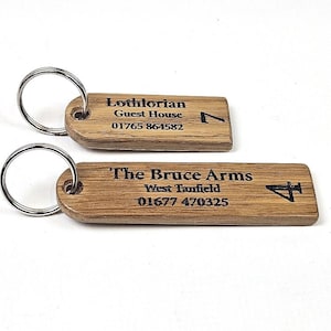 Puede incluir: Dos llaveros rectangulares de madera con anillas metálicas. El llavero superior está grabado con Lothlorian Guest House, 01765 864582 y el número 7. El llavero inferior está grabado con The Bruce Arms, West Tanfield, 01677 470325 y el número 4.