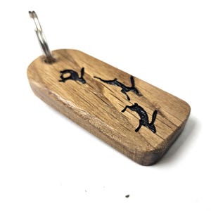 Puede incluir: Un llavero de madera con un anillo plateado. La etiqueta rectangular presenta tres siluetas negras de liebres saltando. La veta de la madera es visible y el llavero está sobre un fondo blanco.