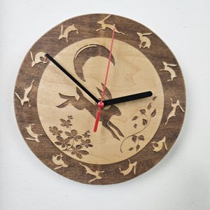May include: A round wooden clock with a light wood center and a darker wood border. The clock face features a laser-cut design of a goat, rabbits, and floral elements. The clock hands are black with a red second hand.