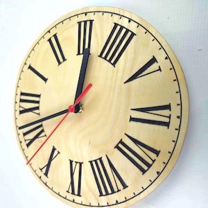 May include: A round wooden wall clock with a light natural wood grain finish. The clock face features bold black Roman numerals, two black hour and minute hands, and a thin red second hand pointing toward the lower left quadrant.