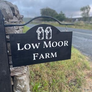 Może przedstawiać: Czarny drewniany szyld z białym napisem "Low Moor Farm" i ilustracjami baniek na mleko. Szyld wisi na czarnym metalowym wsporniku na kamiennym murze, na tle wiejskiego krajobrazu.