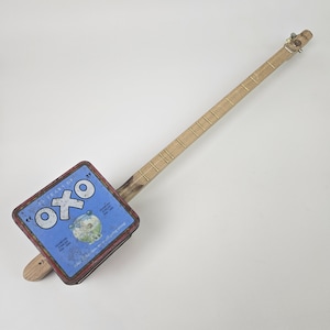 May include: A handcrafted cigar box guitar featuring an OXO tin box in blue and red as its body. The neck is made of light wood, with metal frets and tuning pegs. A unique, rustic musical instrument.