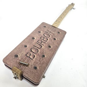 Könnte beinhalten: Eine einzigartige Zigarrenkasten-Gitarre mit einem braunen, rechteckigen Korpus, der einem Keks ähnelt und das Wort "BOURBON" trägt. Sie hat einen Holz-Hals, Saiten und Metallbeschläge.