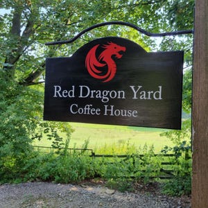 May include: A black wooden sign with the words "Red Dragon Yard Coffee House" in white lettering. A red dragon logo is at the top. The sign is hanging outside, surrounded by green foliage and a field in the background.