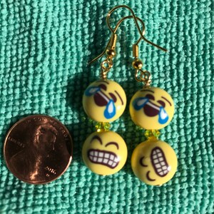May include: A pair of gold dangle earrings with yellow and brown emoji beads. The top bead is a yellow emoji with a tear rolling down its cheek. The bottom bead is a yellow emoji with a wide open mouth and teeth showing. The earrings are about the size of a US penny.