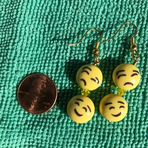May include: A pair of gold dangle earrings with yellow smiley face charms. The charms have different expressions, including winking and a neutral expression.