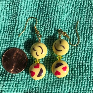 May include: A pair of yellow emoji earrings with gold hooks. One earring has a smiley face with closed eyes and the other has a smiley face with hearts for eyes.