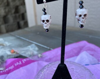 May include: A pair of silver skull earrings with black crystal beads. The skulls are white with black eyes and are hanging from silver hooks.