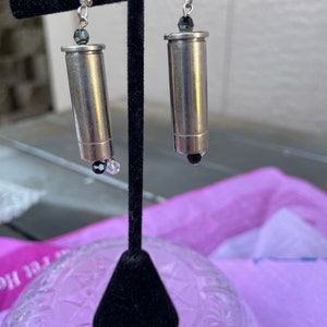 May include: A pair of silver bullet earrings with black beads hanging from the bottom. The earrings are displayed on a black stand.