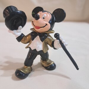 May include: A black and gold plastic figurine of Mickey Mouse wearing a tuxedo and holding a top hat and a cane.