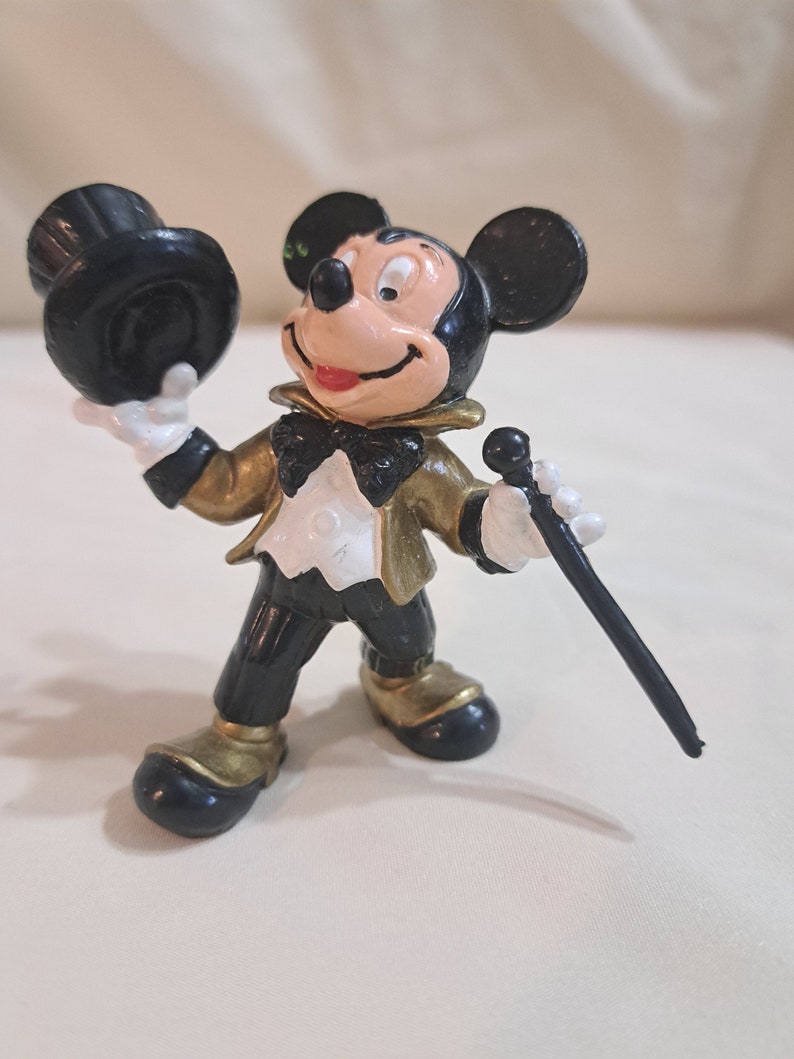 May include: A black and gold plastic figurine of Mickey Mouse wearing a tuxedo and holding a top hat and a cane.
