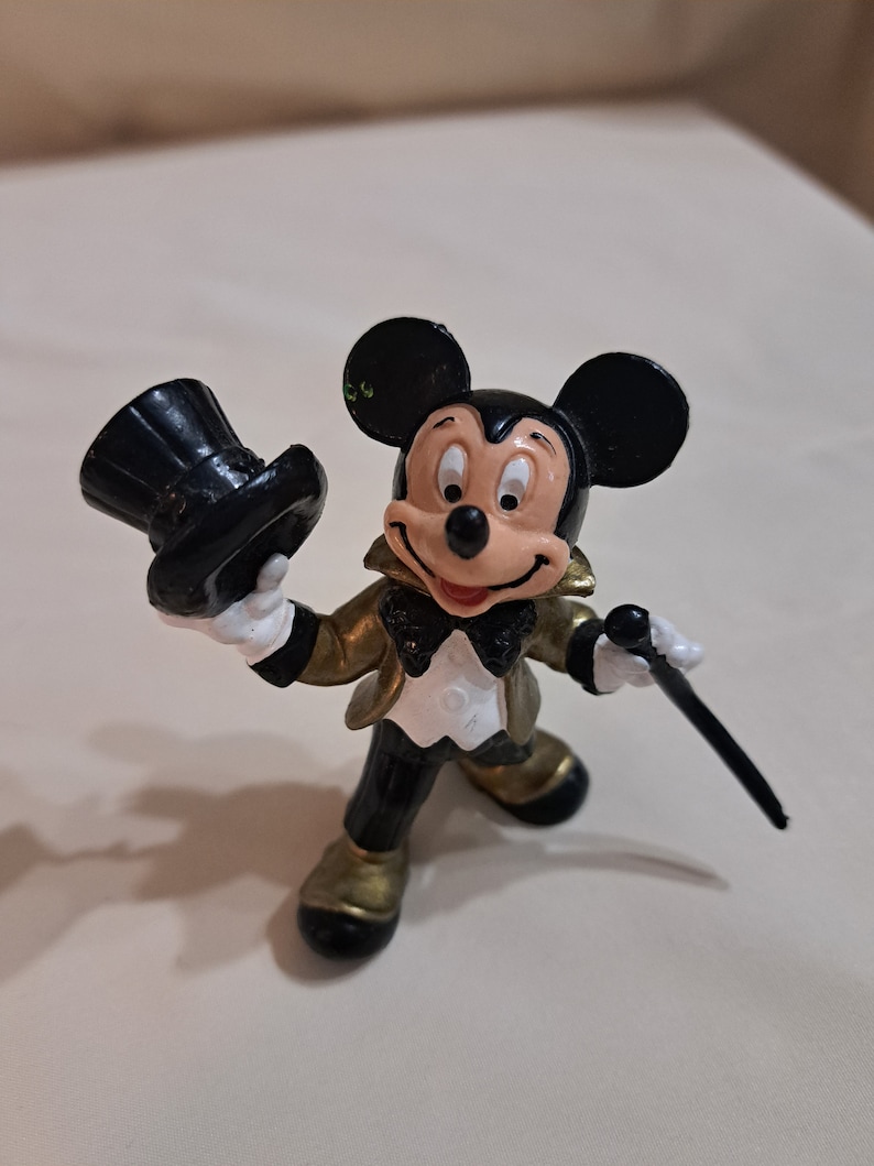 May include: A black and gold Mickey Mouse figurine wearing a black top hat and holding a black cane. The figurine is in a formal outfit with a white bow tie and a gold jacket.