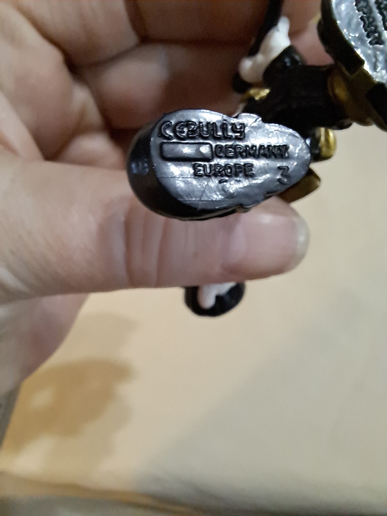 May include: Close-up of a black plastic toy figure with a white and gold design. The figure has a label on the bottom that reads "CEBULY GERMANY EUROPE 3".