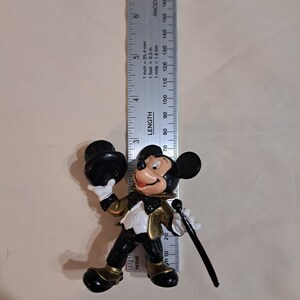 May include: A black and gold plastic figurine of Mickey Mouse wearing a tuxedo and holding a top hat and a cane. The figurine is approximately 19 cm tall.