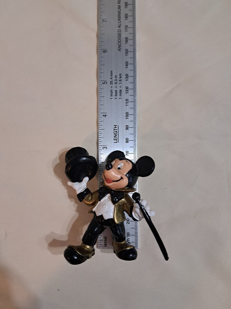 May include: A black and gold plastic figurine of Mickey Mouse wearing a tuxedo and holding a top hat and a cane. The figurine is approximately 19 cm tall.