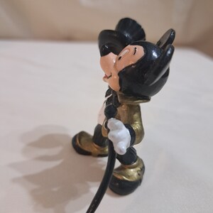 May include: A black and gold Mickey Mouse figurine with a black top hat and a cane. The figurine is standing on a white surface.