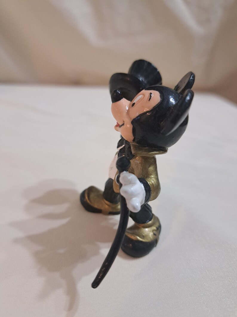 May include: A black and gold Mickey Mouse figurine with a black top hat and a cane. The figurine is standing on a white surface.