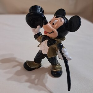 May include: A black and gold Mickey Mouse figurine wearing a black top hat and holding a black cane. The figurine is in a standing pose with a smile on its face.