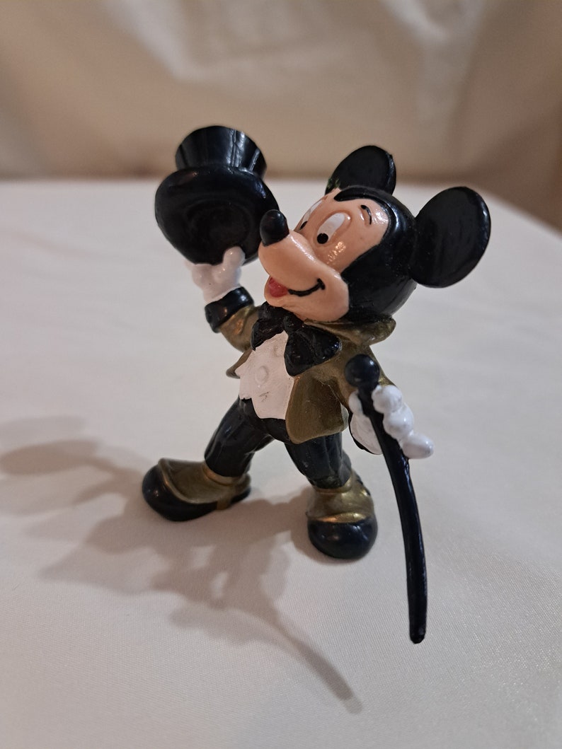 May include: A black and gold Mickey Mouse figurine wearing a black top hat and holding a black cane. The figurine is in a standing pose with a smile on its face.