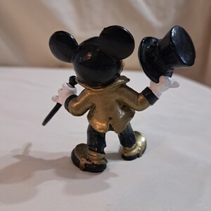 May include: A black and gold Mickey Mouse figurine wearing a black top hat and holding a black cane. The figurine is standing on a white surface.
