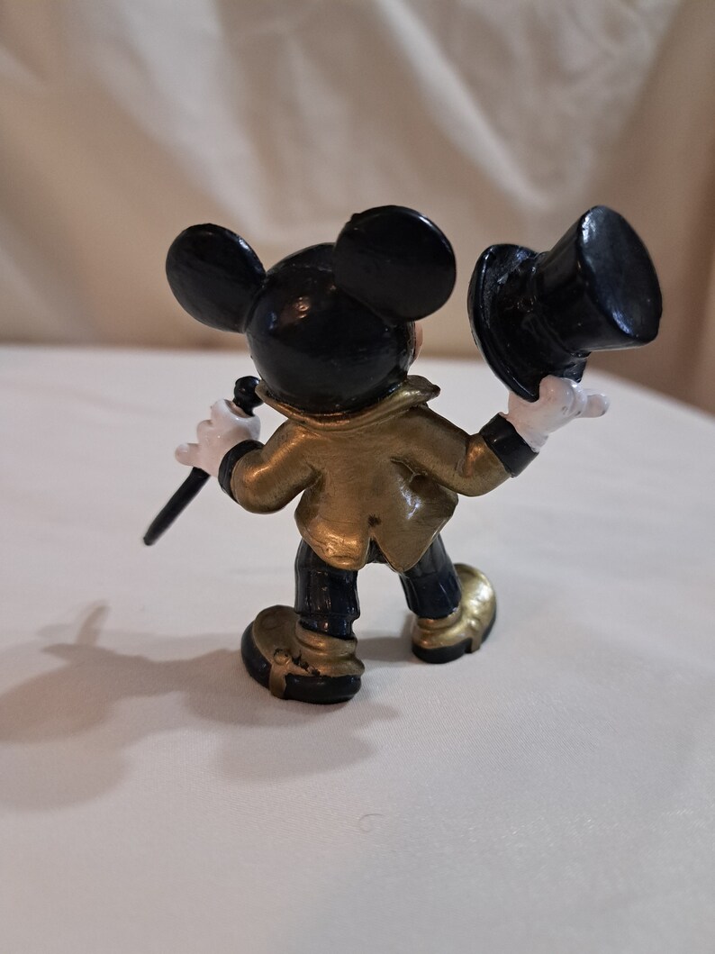 May include: A black and gold Mickey Mouse figurine wearing a black top hat and holding a black cane. The figurine is standing on a white surface.