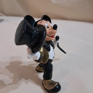 May include: A black and gold Mickey Mouse figurine wearing a tuxedo and holding a top hat. The figurine is made of plastic and is about 4 inches tall.