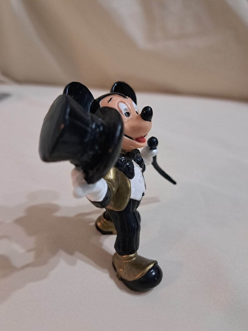 May include: A black and gold Mickey Mouse figurine wearing a tuxedo and holding a top hat. The figurine is made of plastic and is about 4 inches tall.