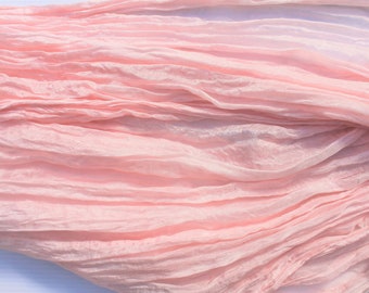 May include: A length of pale pink fabric with a crinkled texture. The material is arranged in soft folds, creating a layered effect. The colour shifts from a deeper pink to a lighter shade, suggesting a gradient.
