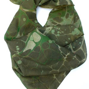 May include: A green scarf with a marbled design in shades of olive and white. The scarf is knotted, displaying the pattern. The fabric looks lightweight and flowing, suitable for different styles. The scarf is made of a soft material.