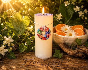 May include: A lit white pillar candle with a colorful circular design, tied with string. The candle sits on a wooden surface with the words "Free Spirit" and is surrounded by greenery, flowers, and a bowl of orange slices.